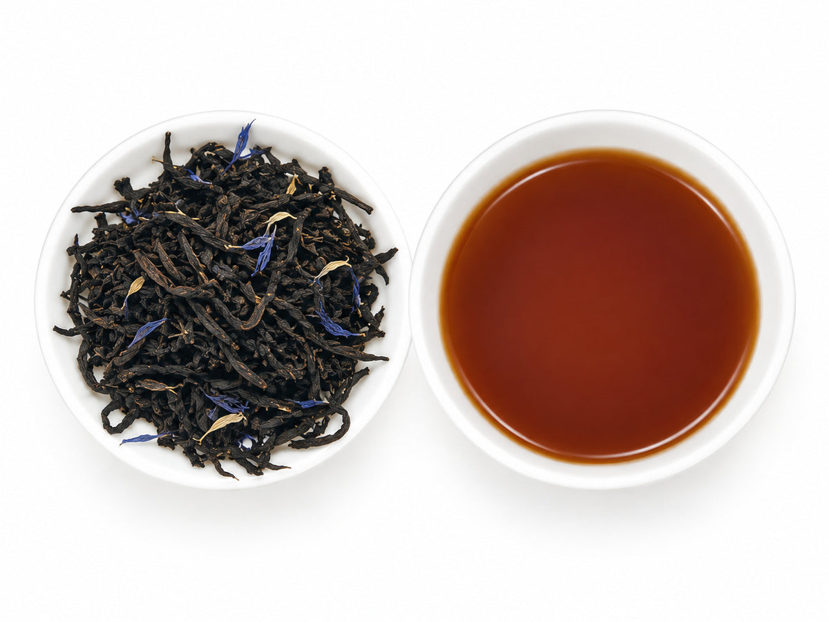 Earl Grey Blue Clue, aromatisierter Schwarztee mit dunklen Teeblättern und blauen Blüten in einer weißen Schale, daneben eine weiße Teeschale mit braunem Aufguss.