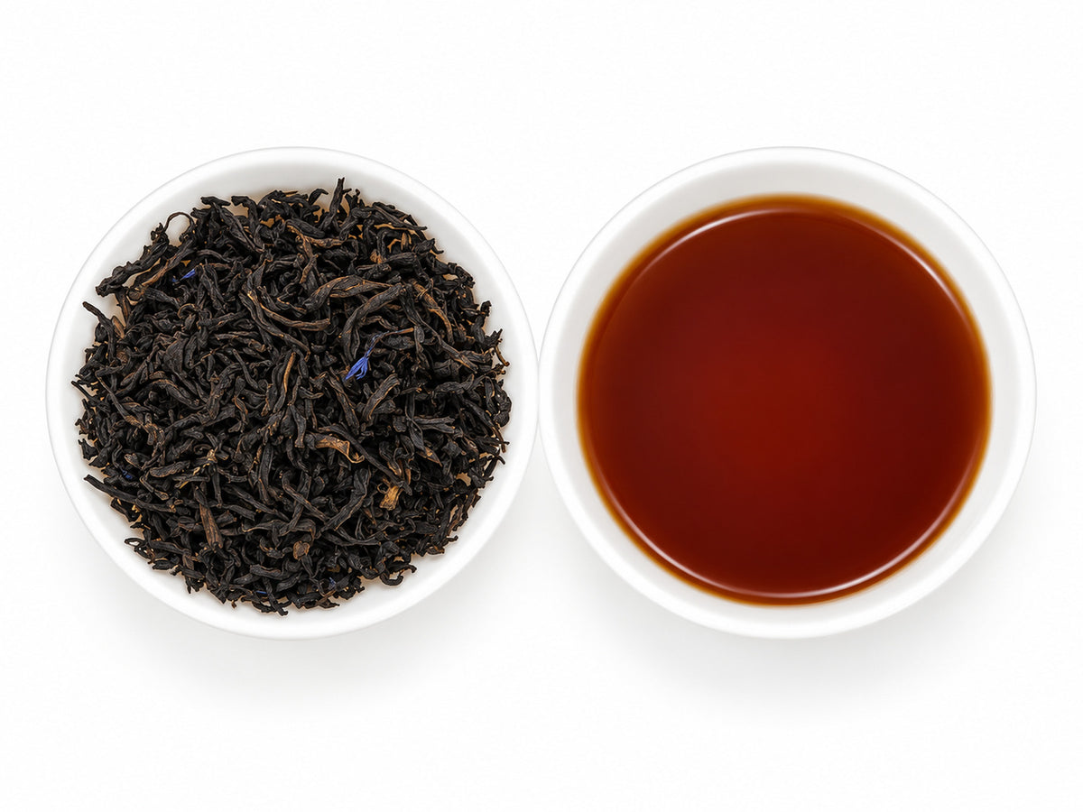 Earl Grey Lady Grey, aromatisierter Schwarztee mit dunklen, gedrehten Teeblättern und einzelnen blauen Blüten, daneben eine weiße Teeschale mit braunem Aufguss.