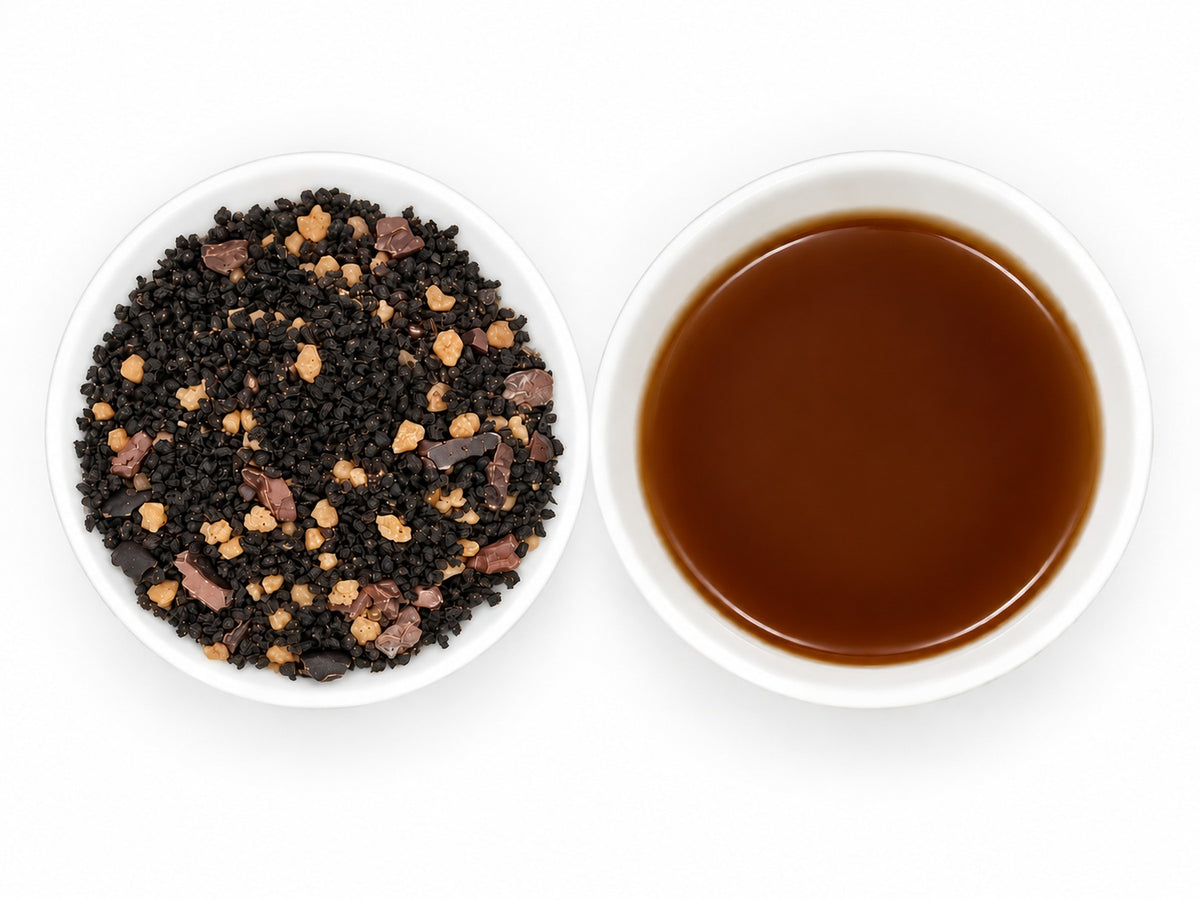 Haselnuss Krokant – aromatisierter Schwarztee mit dunklen, feinen Teeblättern, goldbraunen Krokantstückchen und braunen Stückchen auf einem weißen Tellerchen, daneben eine weiße Teeschale mit klarem, dunkelbraunem Aufguss.
